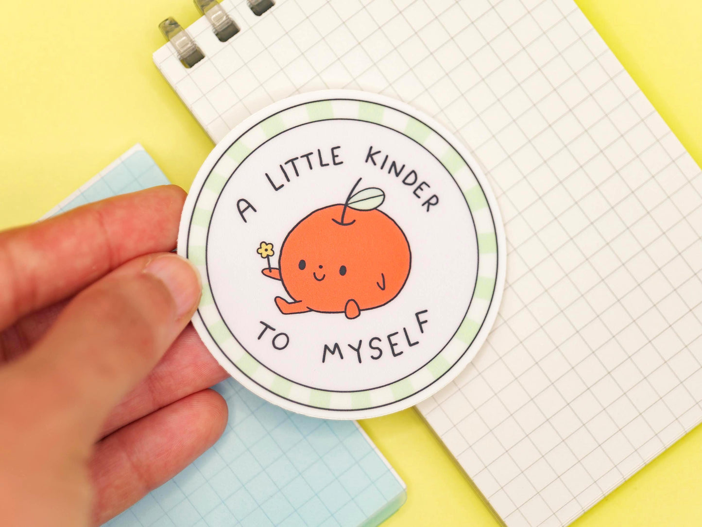 Circular sticker with a cartoon character of an apple and text saying "a little kinder to myself" held by a hand on a yellow background