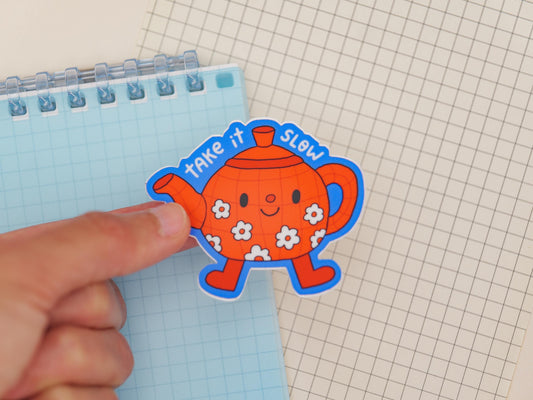 Hand holding a colorful teapot-shaped sticker with text on a grid background