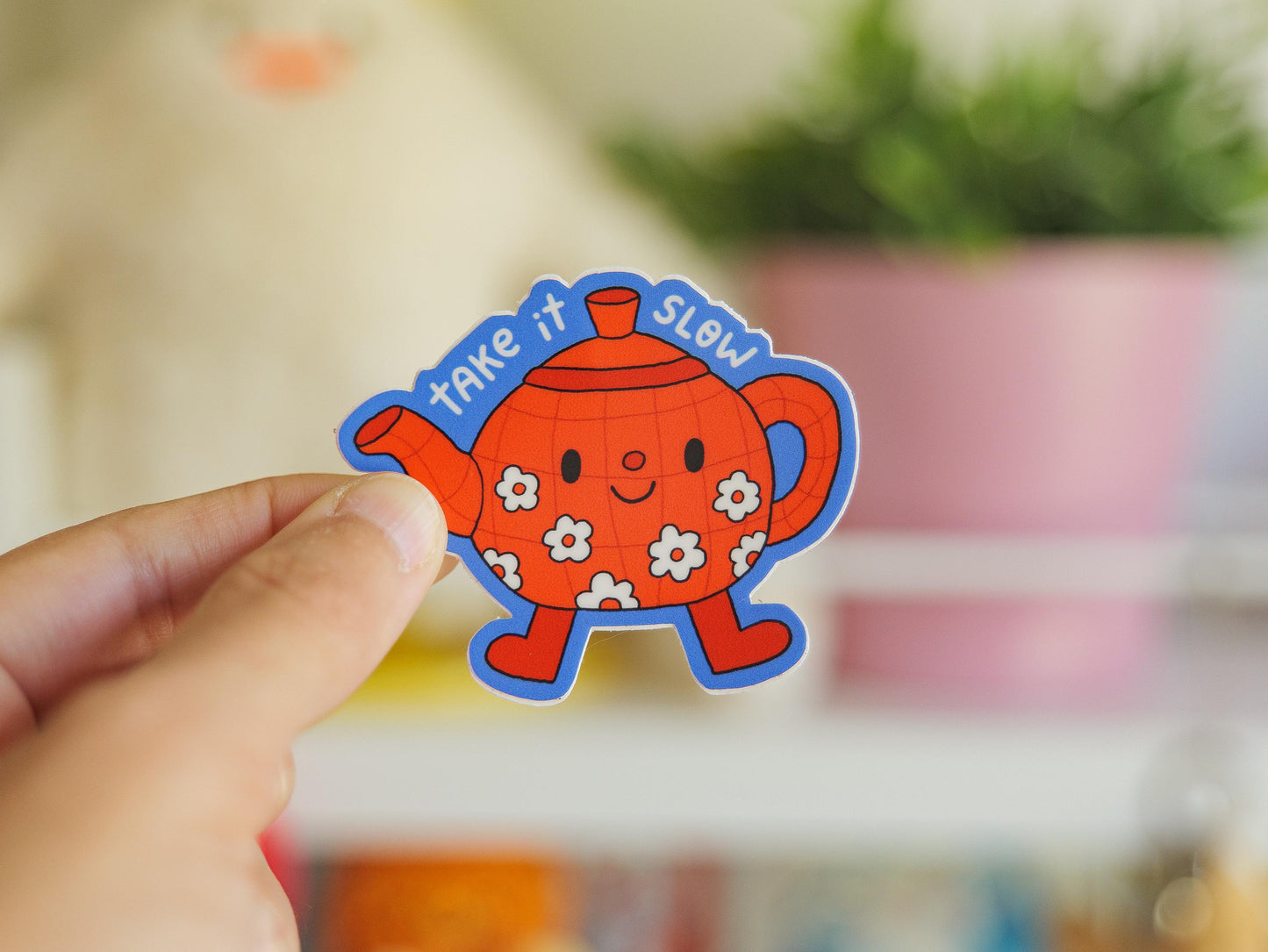 Hand holding a colorful teapot-shaped sticker with text 'take it SLOW' against a blurred indoor background.