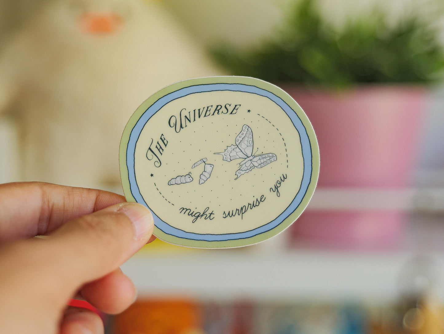 Circular sticker with text saying "the universe might surprise you" and illustrations of a catterpillar turning into a chrysalis and butterfly, held by a hand, blurred background