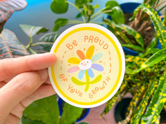 Hand holding a sticker with a flower design and text 'Be proud of your growth' against a plant background.