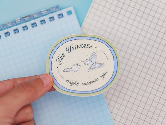 Circular sticker with text saying "the universe might surprise you" and illustrations of a catterpillar turning into a chrysalis and butterfly, on a blue background held over a notebook and grid paper background