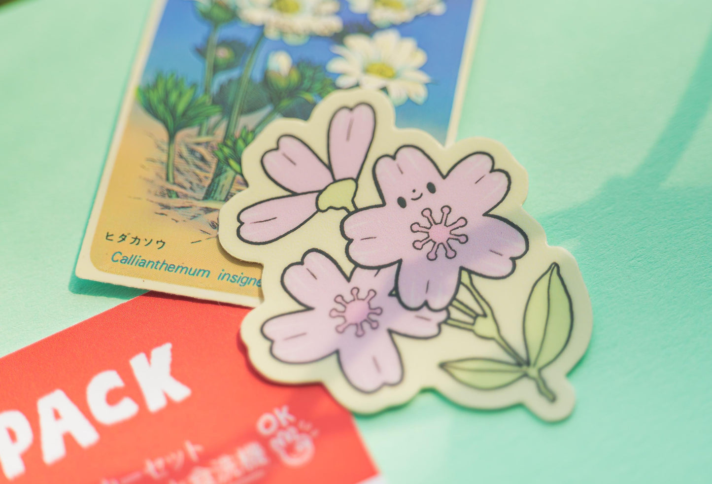 Sakura flower sticker with a smiley face on a green surface with a card and red pack in the background