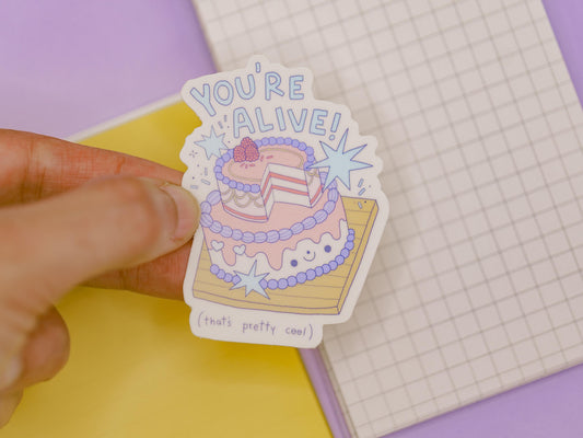 Sticker with a cake design and text held by a hand on a colorful background