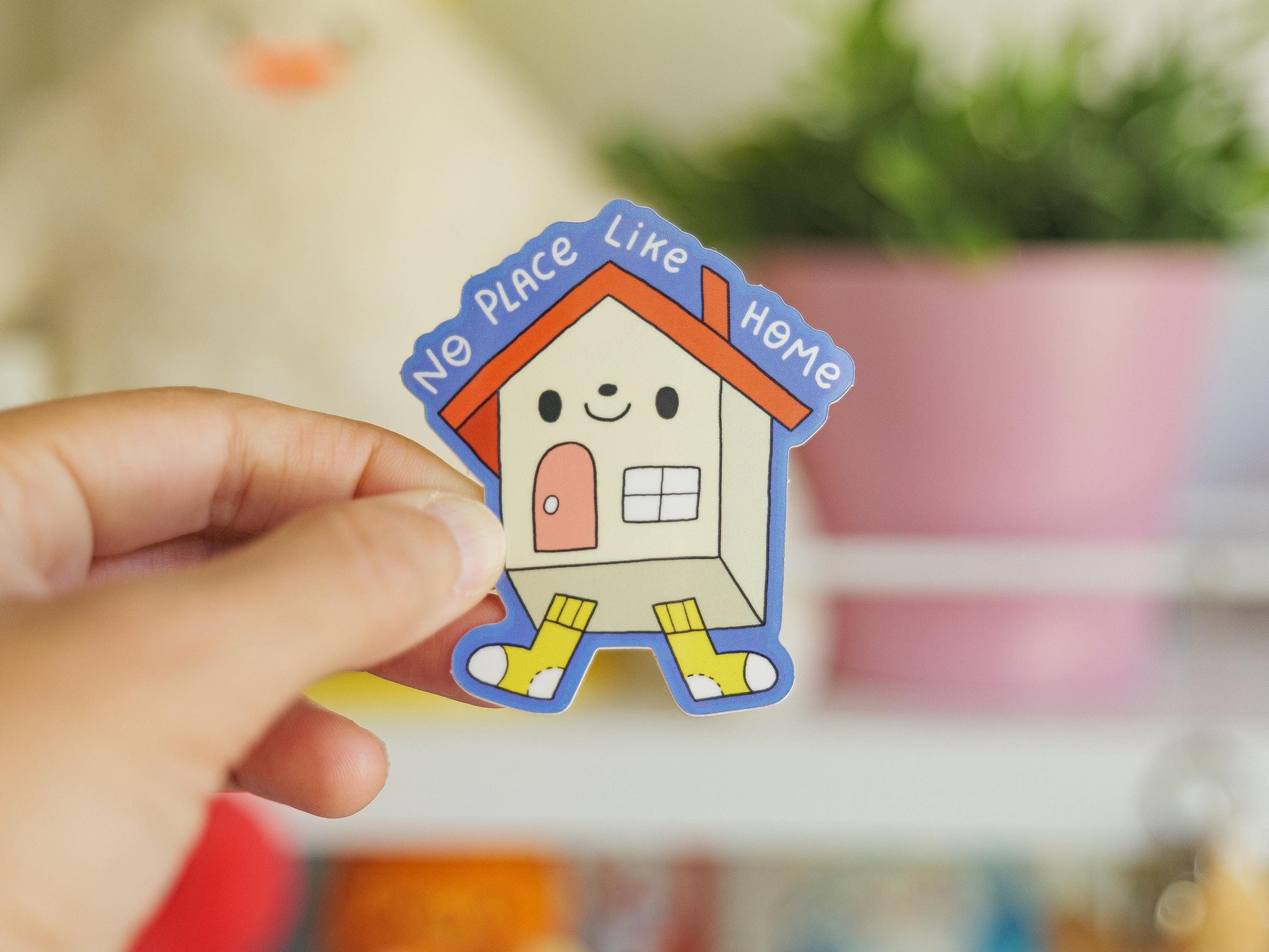 Hand holding a colorful house-shaped sticker with text 'No Place Like Home' against a blurred indoor background.