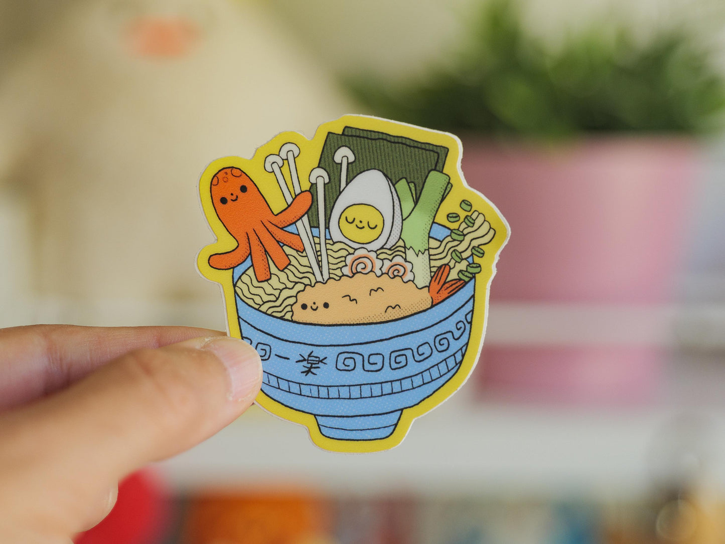 Sticker of a bowl of ramen with cartoon characters held by a hand against a blurred background.