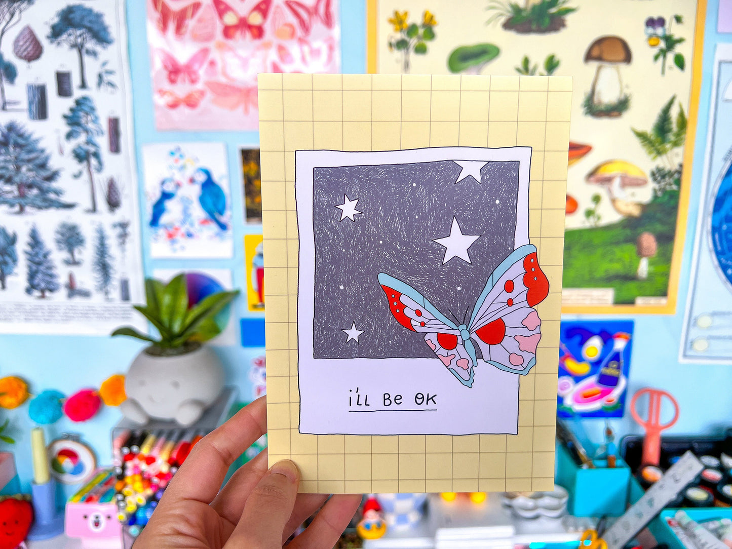 Butterfly illustration on a postcard held in front of a colorful background with various illustrations.