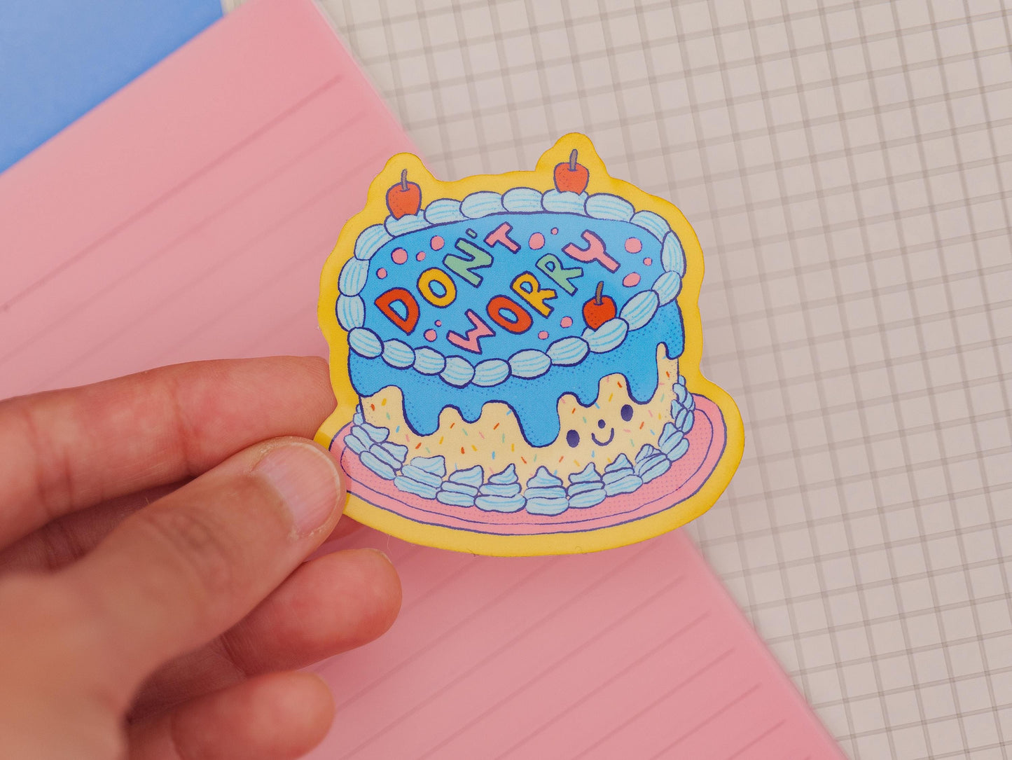 Sticker with a cake design and 'Don't Worry' text held by a hand on a checkered background