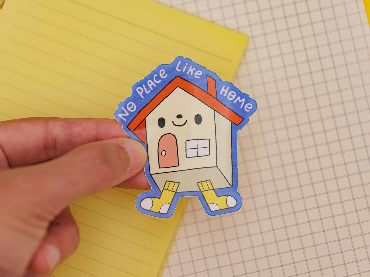 Sticker of a house with 'No Place Like Home' text held over a yellow notepad and grid paper.