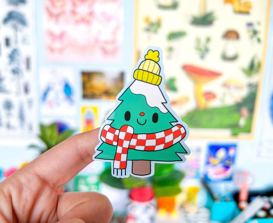 Sticker of a Christmas tree/Balsam Fir with a yellow hat and red scarf held by a hand against a colorful blurred background.