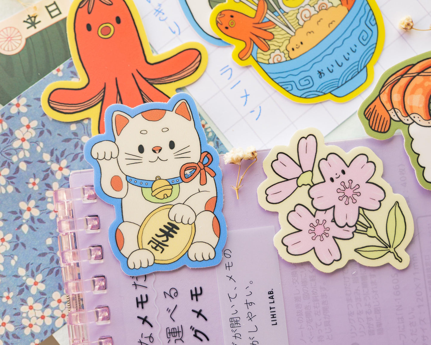 Colorful stickers with various designs on a notebook. Maneki Neko, Daruma, Sakura Flower, Sushi, Onigiri, Tako Sausage, Ramen and Nabe.