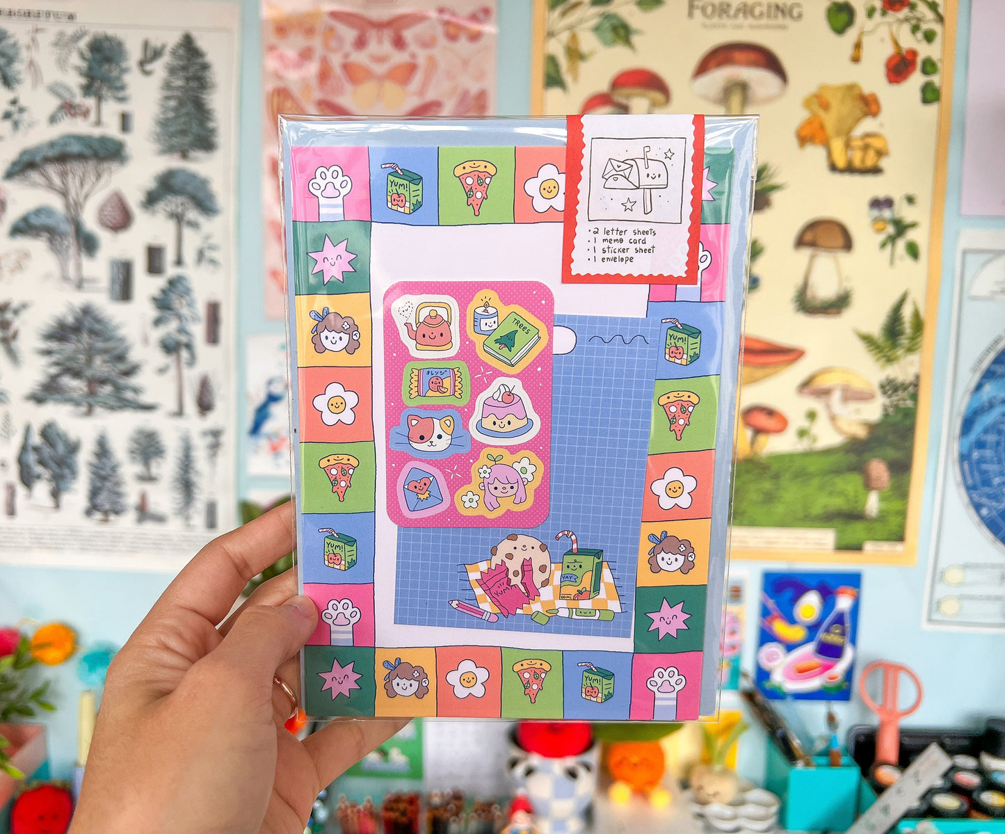 Hand holding a colorful letter writing set with various designs against a background of illustrated posters.