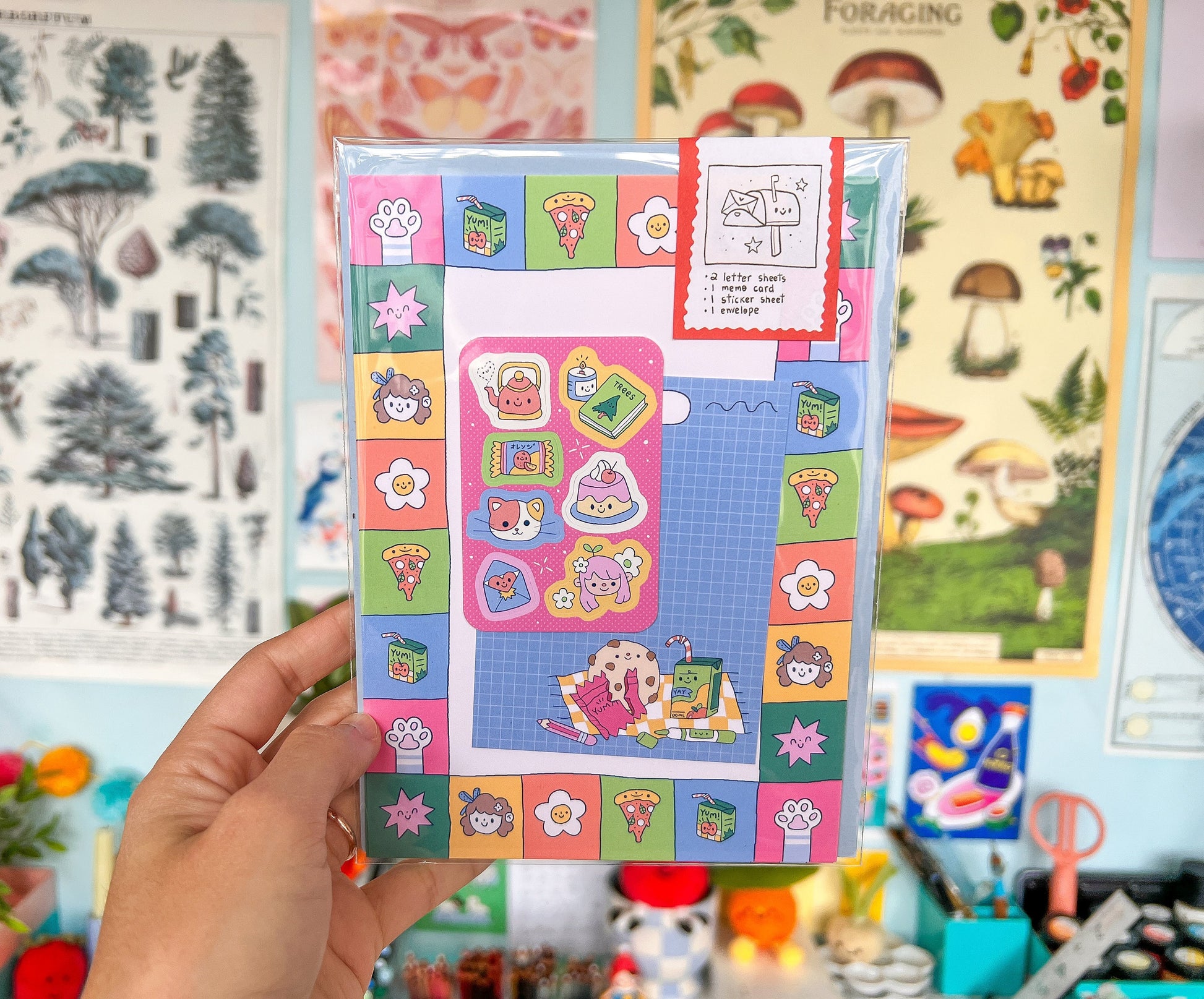 Hand holding a colorful letter writing set with various designs against a background of illustrated posters.