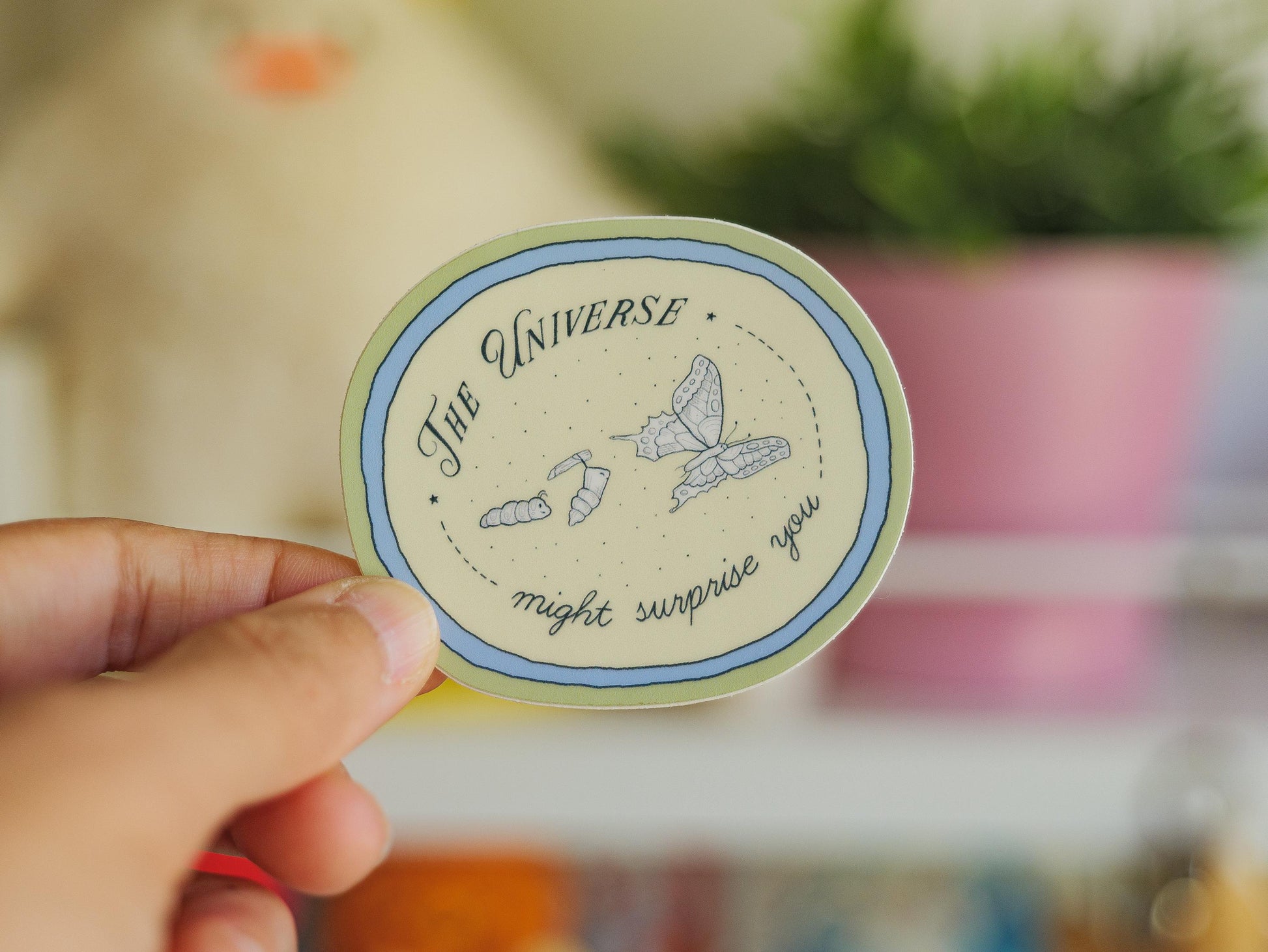 Circular sticker with text saying "the universe might surprise you" and illustrations of a catterpillar turning into a chrysalis and butterfly, held by a hand, blurred background