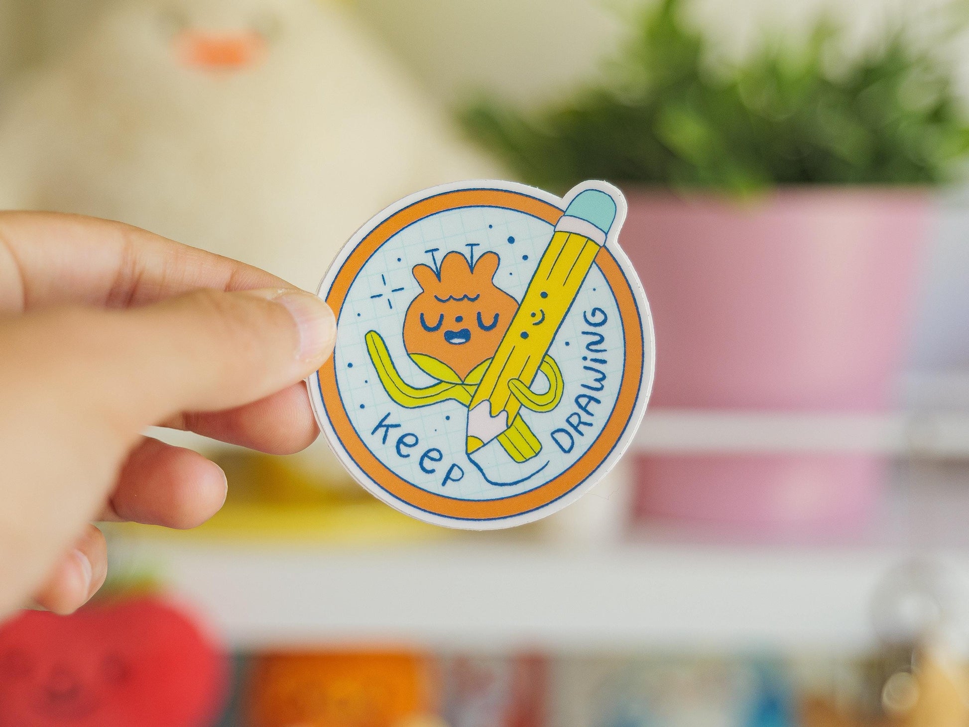 Circular sticker with cartoon character of a flower holding a pencil and text 'keep drawing', held by a hand with a blurred background.