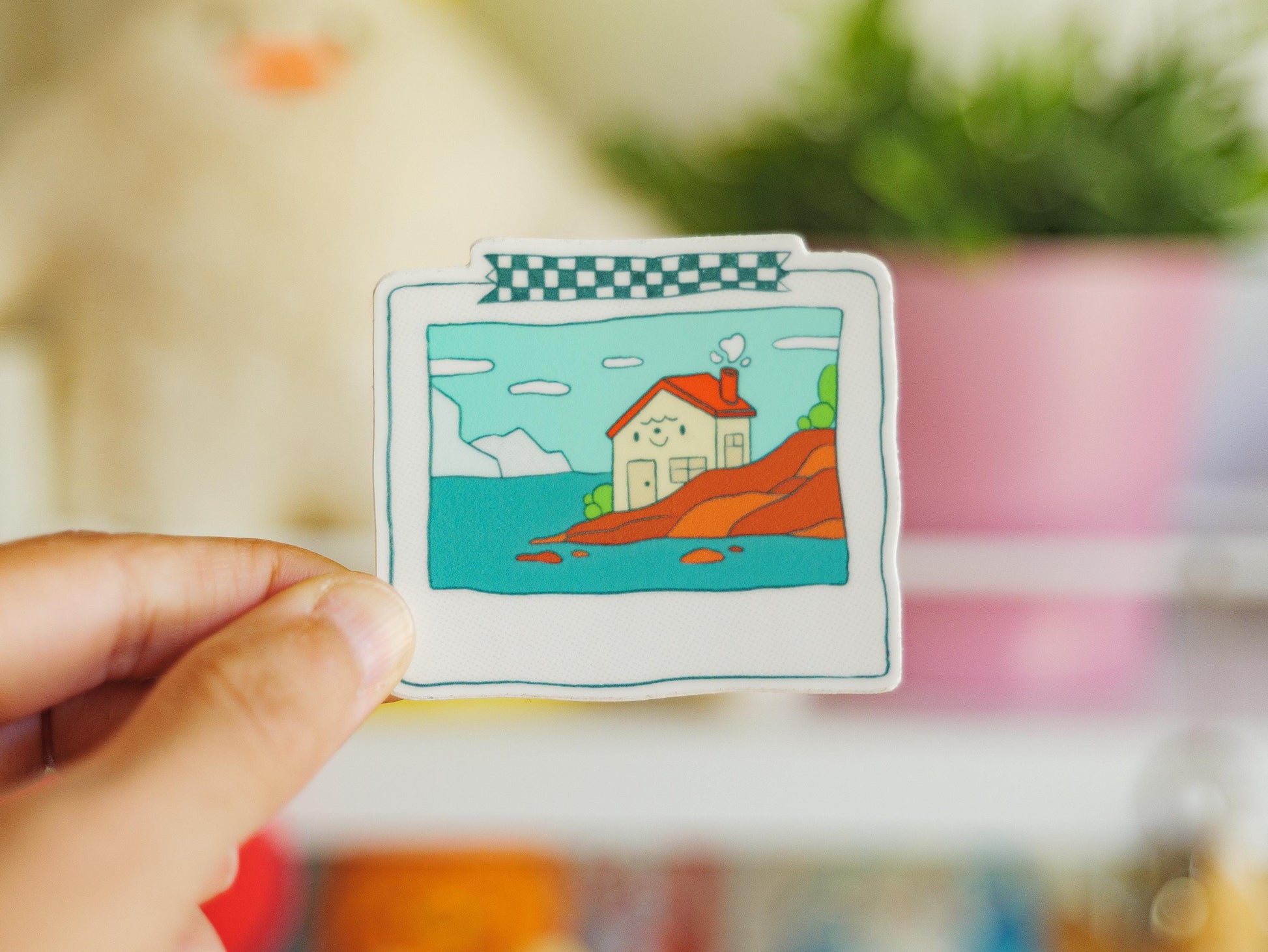 Hand holding a small illustration of a house by a lake with a colorful border.