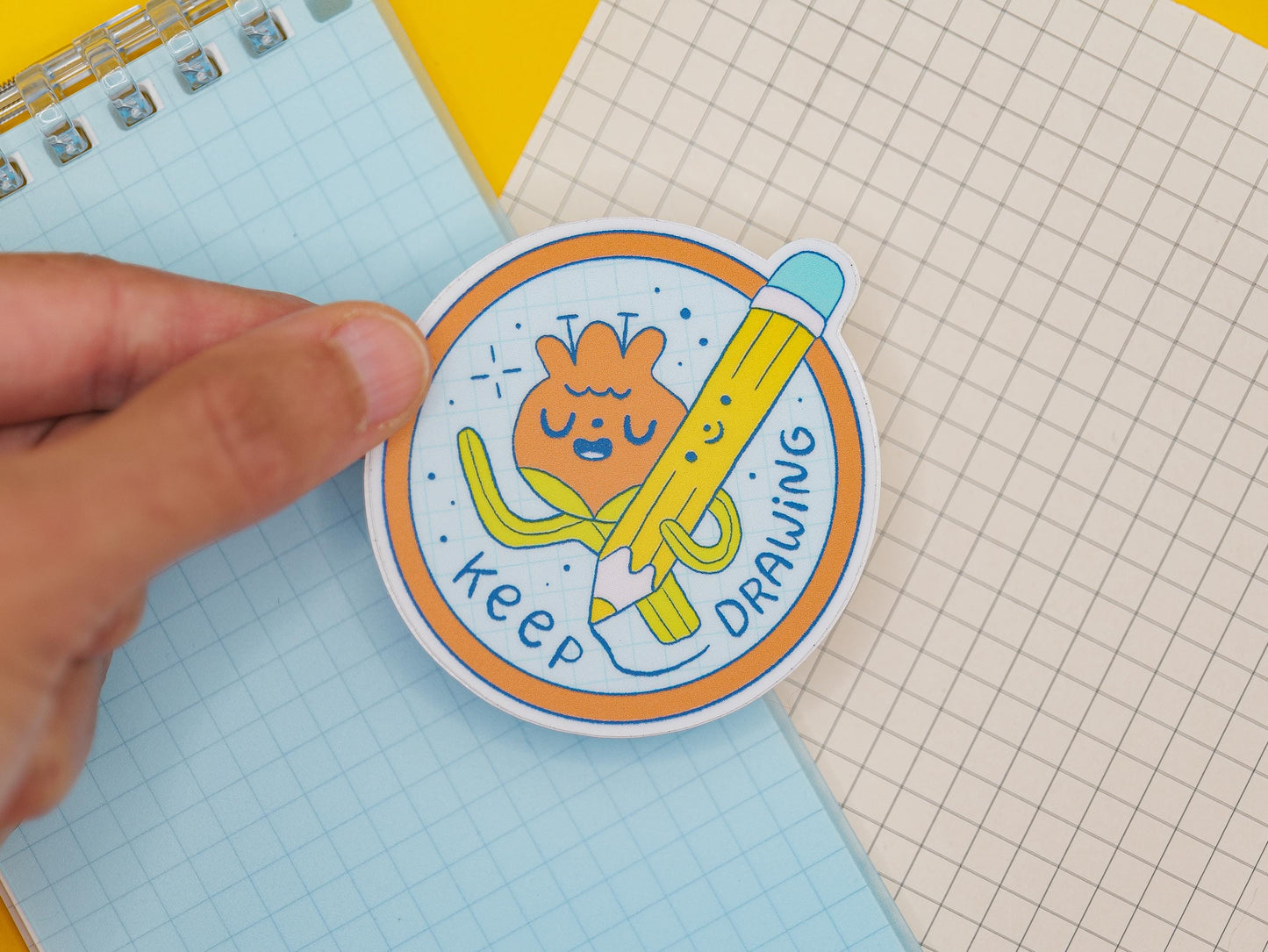 Circular sticker with cartoon character of a flower holding a pencil and text 'keep drawing' on a notebook
