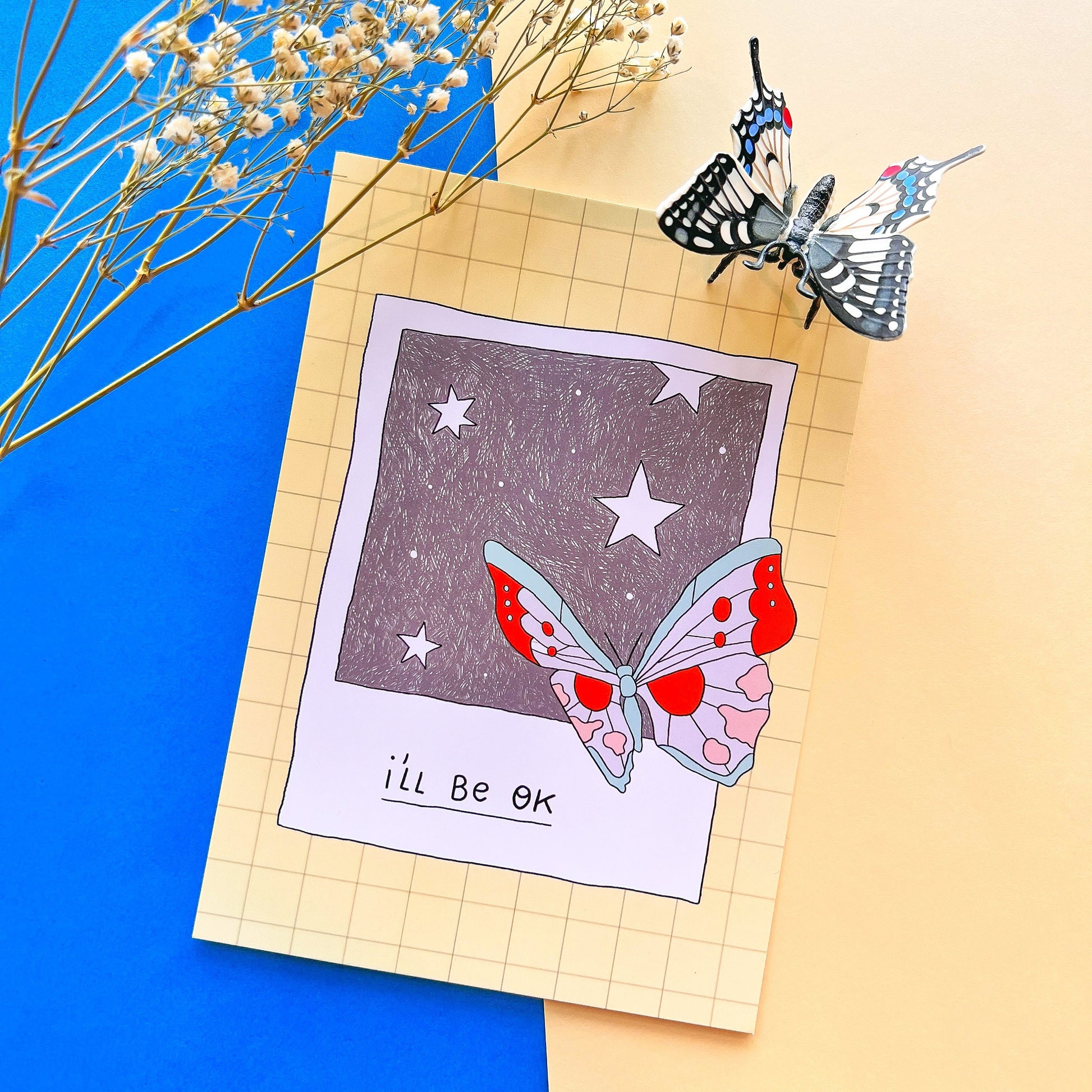 Butterfly illustration on a piece of paper with 'I'll Be OK' text, surrounded by dried flowers on a blue and beige background.
