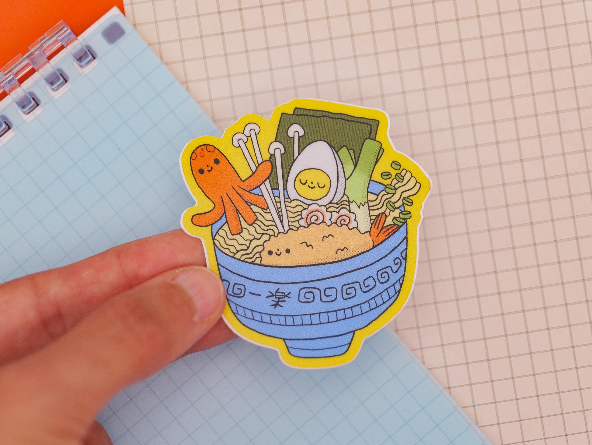 Sticker of a bowl of ramen with an octopus and other toppings held over a notebook.