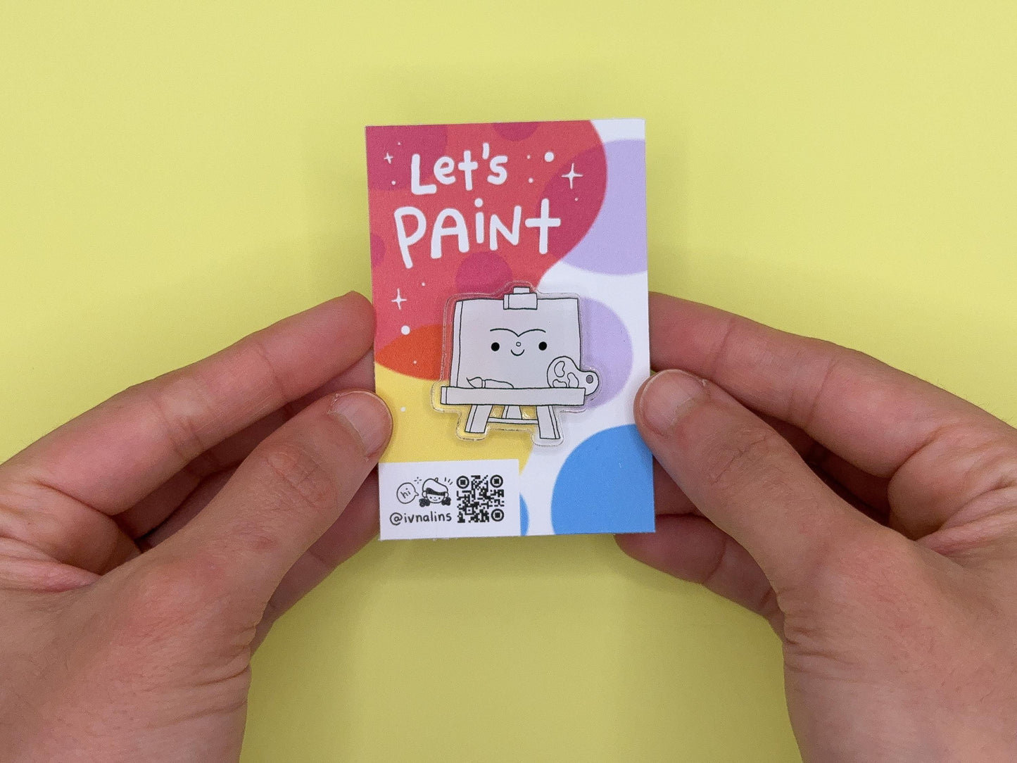 Small colorful card with 'Let's Paint' text, with a pin of a smiling painting easel design, held between two hands against a yellow background