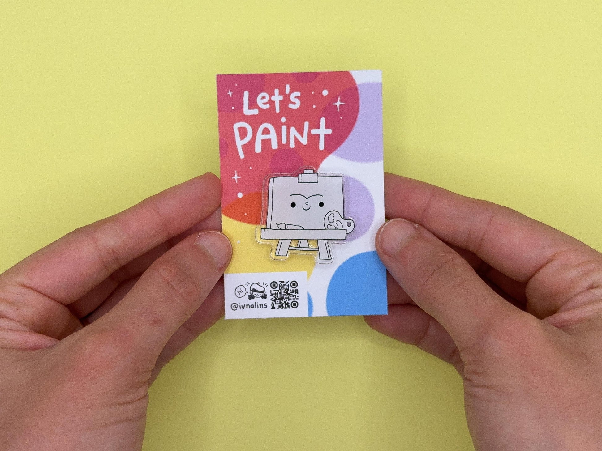Small colorful card with 'Let's Paint' text, with a pin of a smiling painting easel design, held between two hands against a yellow background