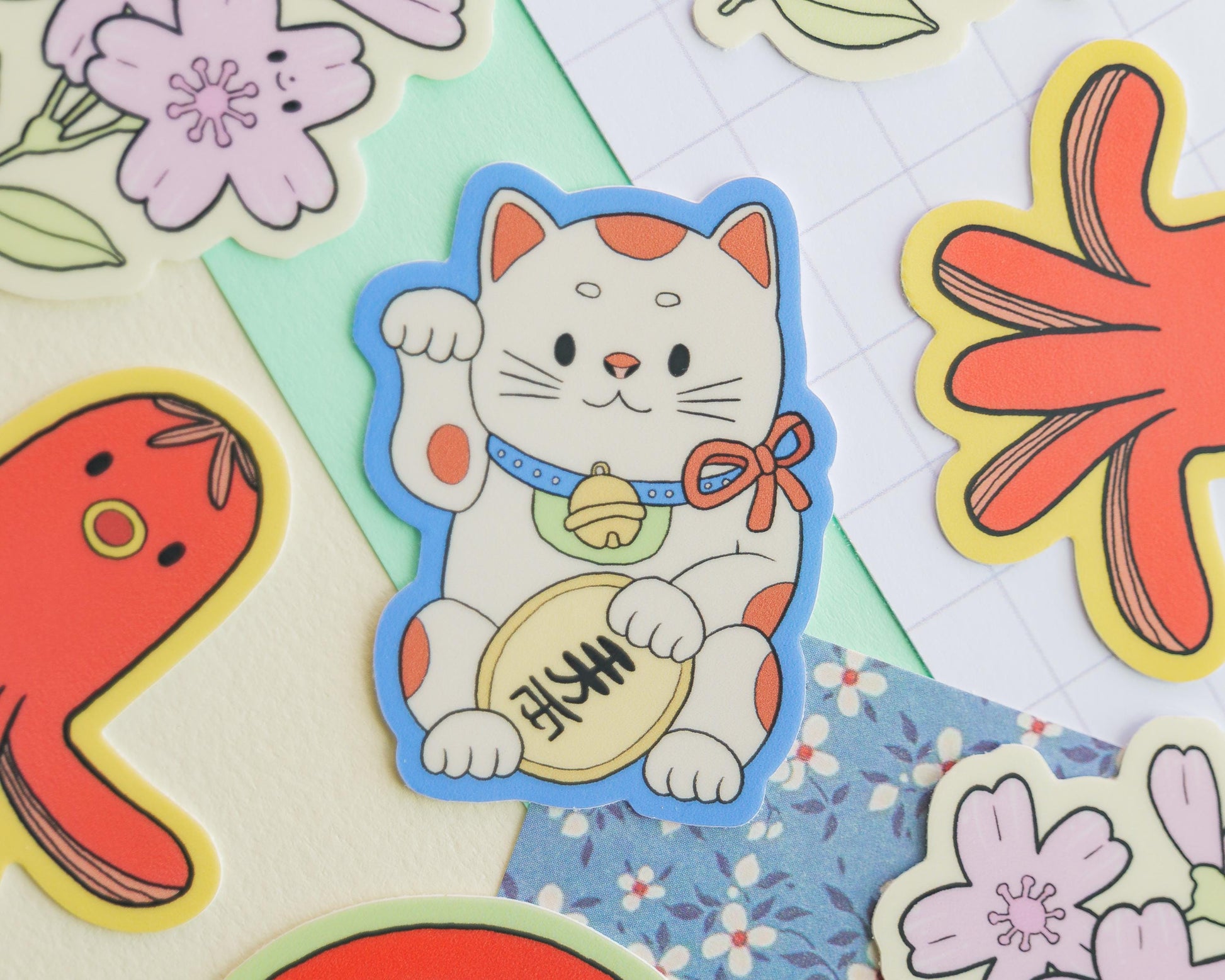 Sticker of a maneki neko cat with a bell, surrounded by colorful flowers and leaves on a textured fabric background.