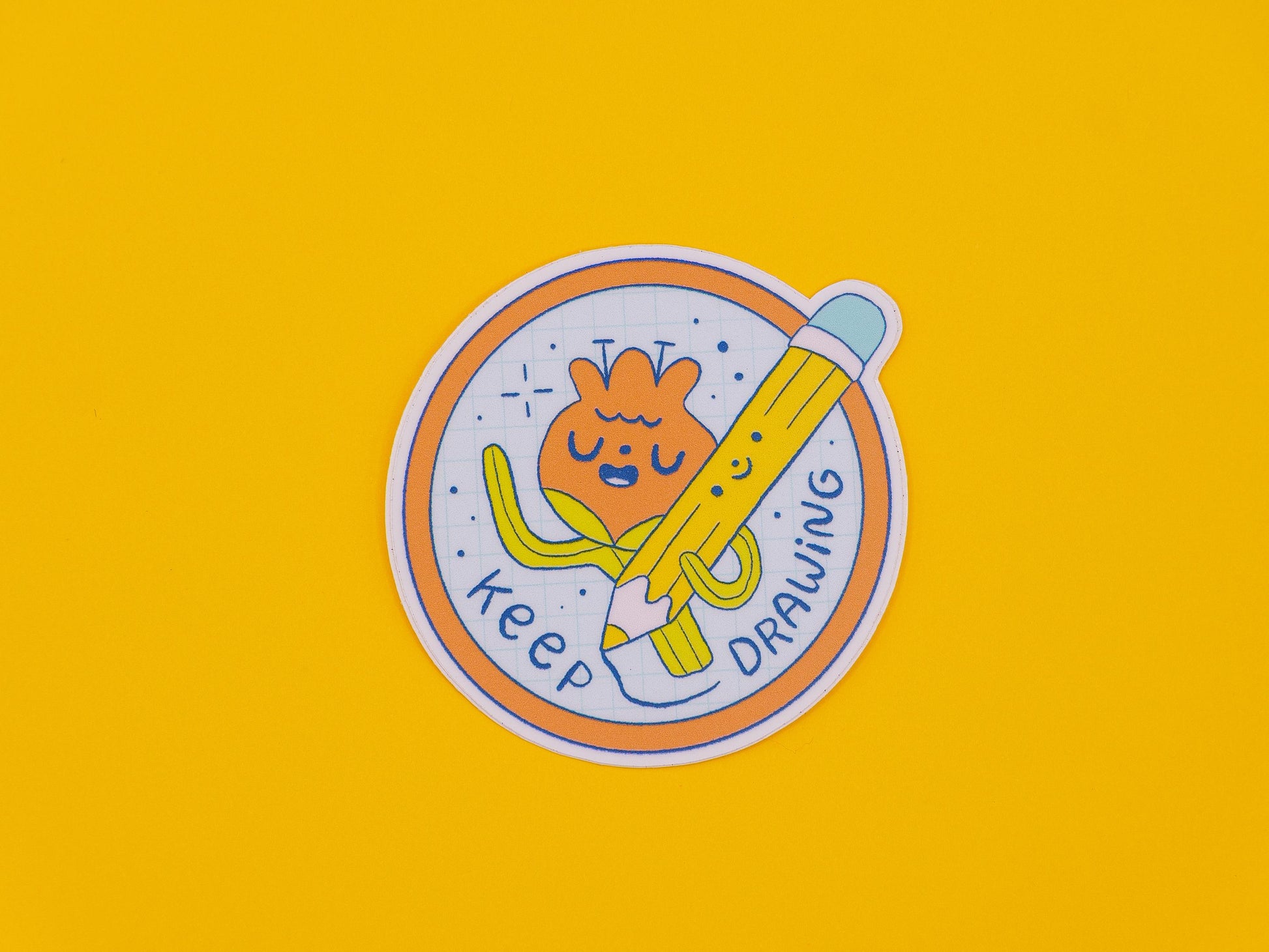 Circular sticker with cartoon character of a flower holding a pencil and text 'keep drawing' on a yellow background