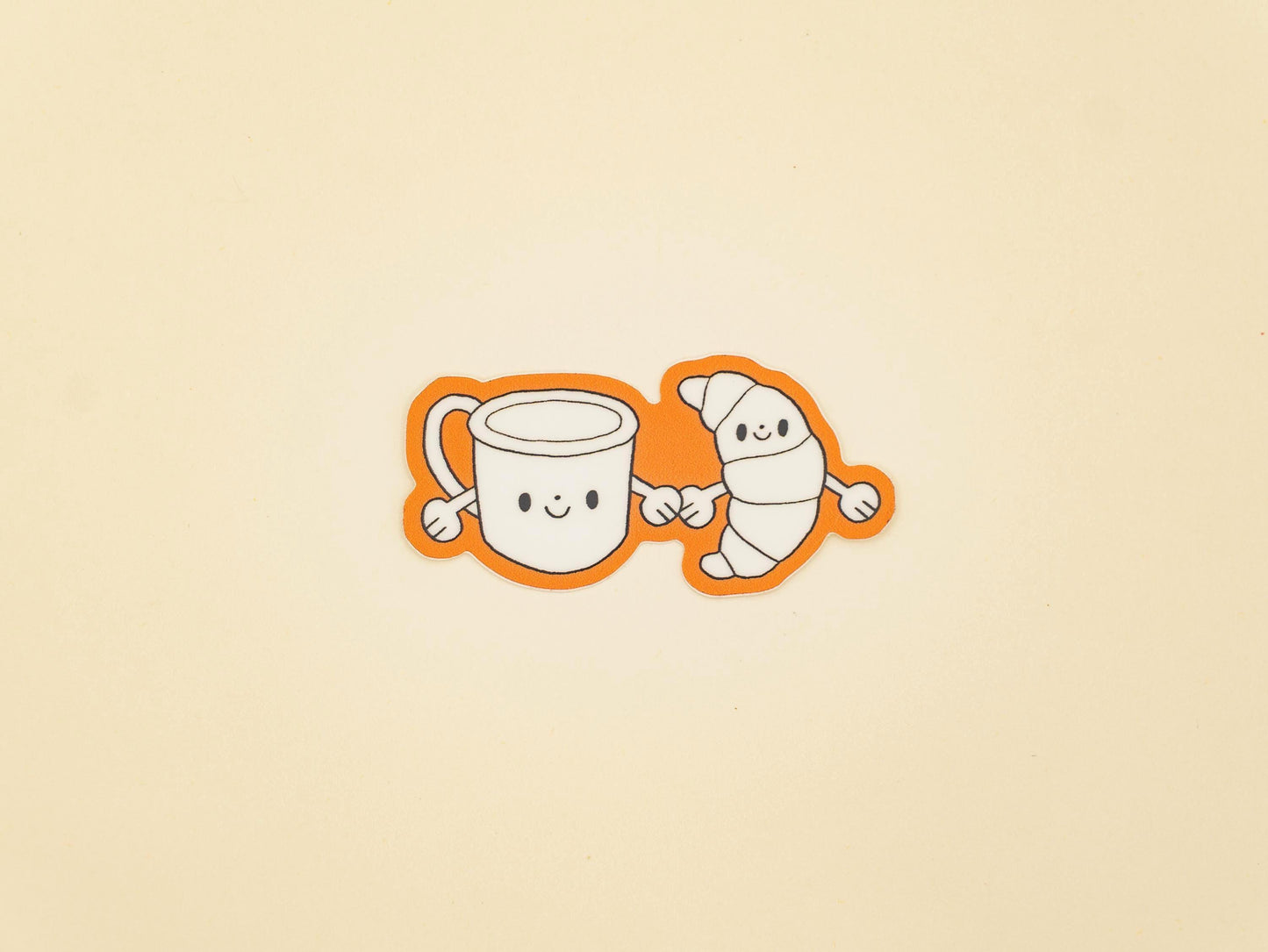 Two cartoon characters, a mug and a croissant, holding hands on a beige background.
