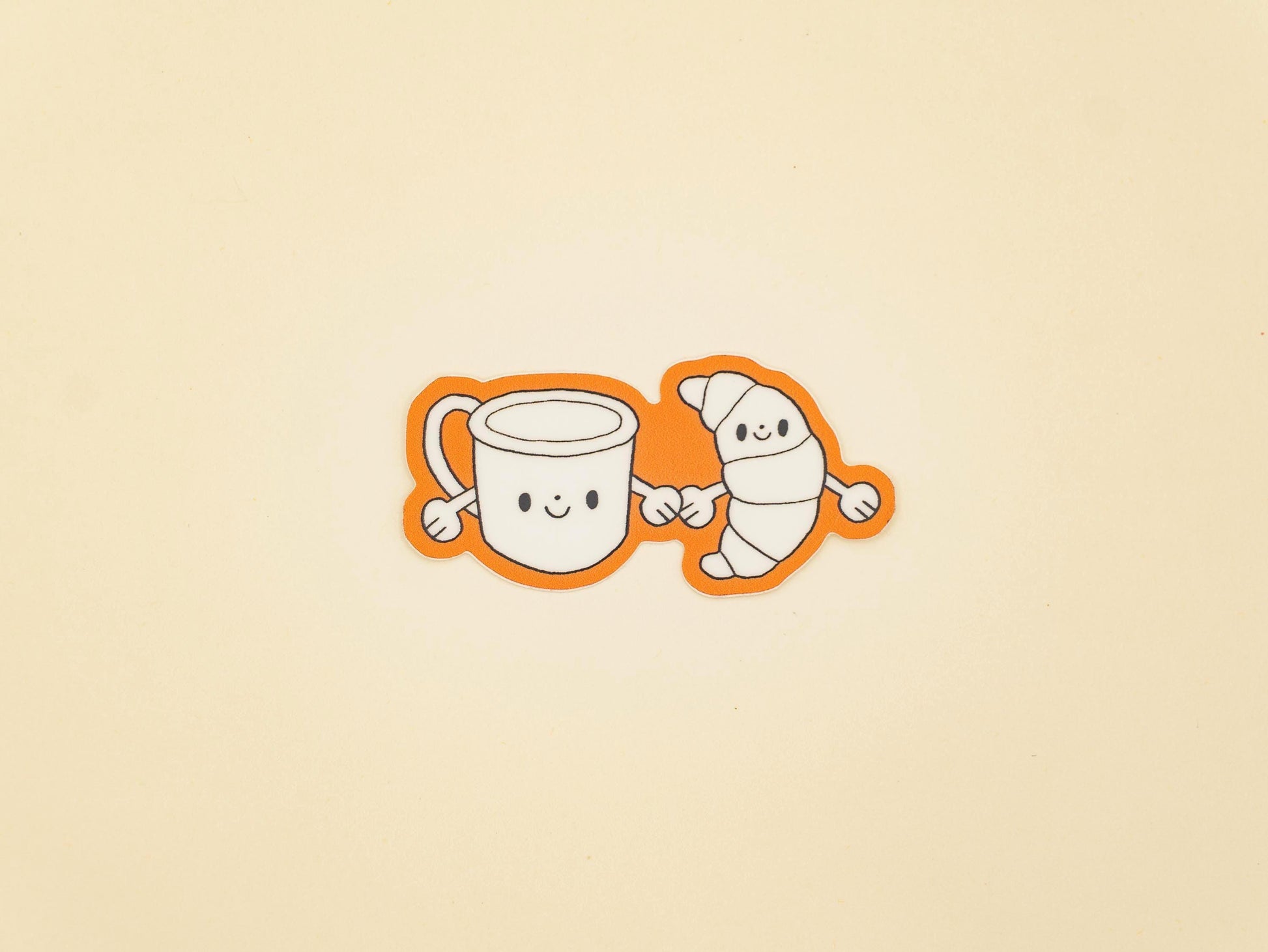 Two cartoon characters, a mug and a croissant, holding hands on a beige background.