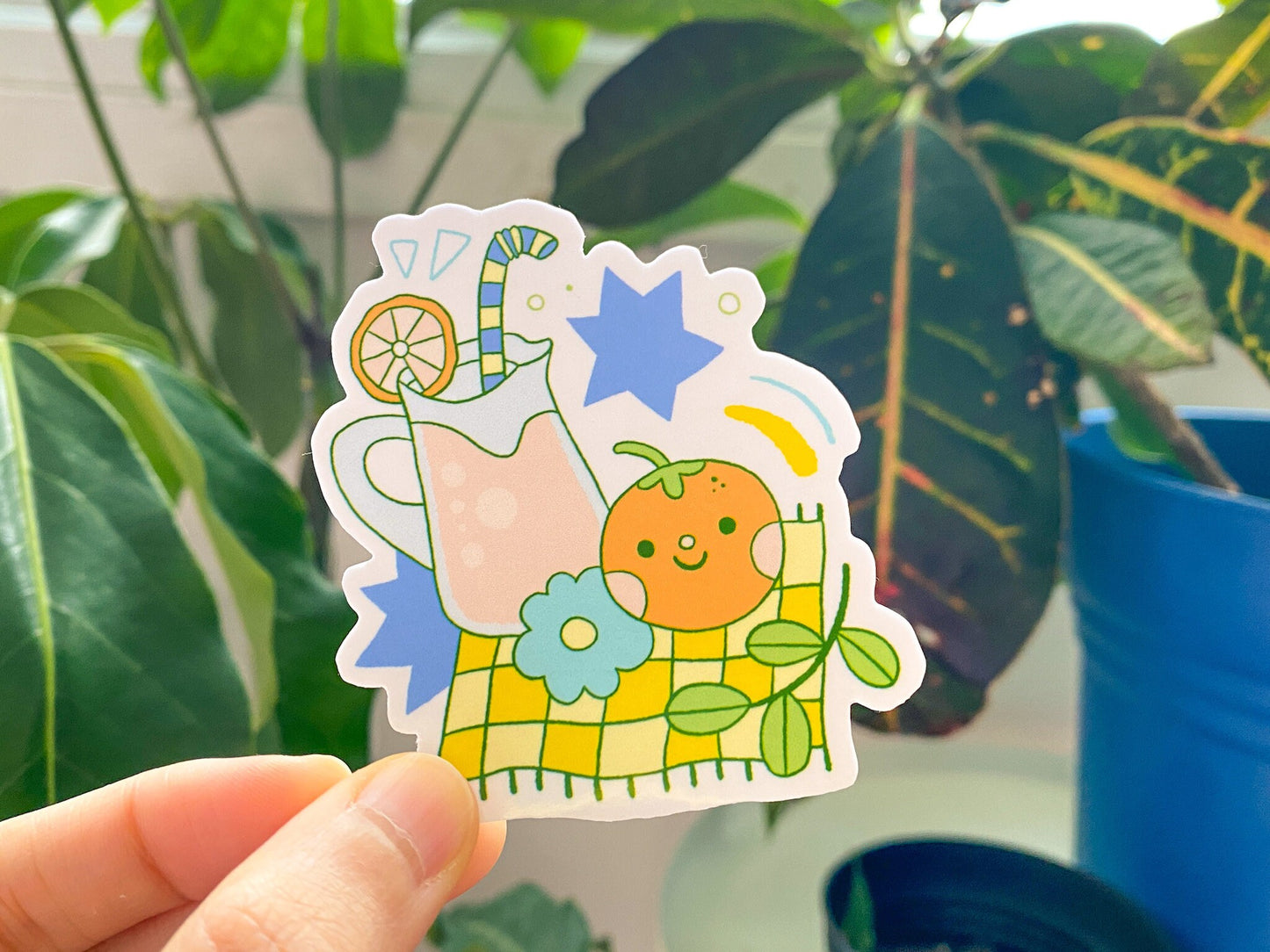 Sticker of a pitcher with lemonade, orange, and flowers held in front of green leaves.