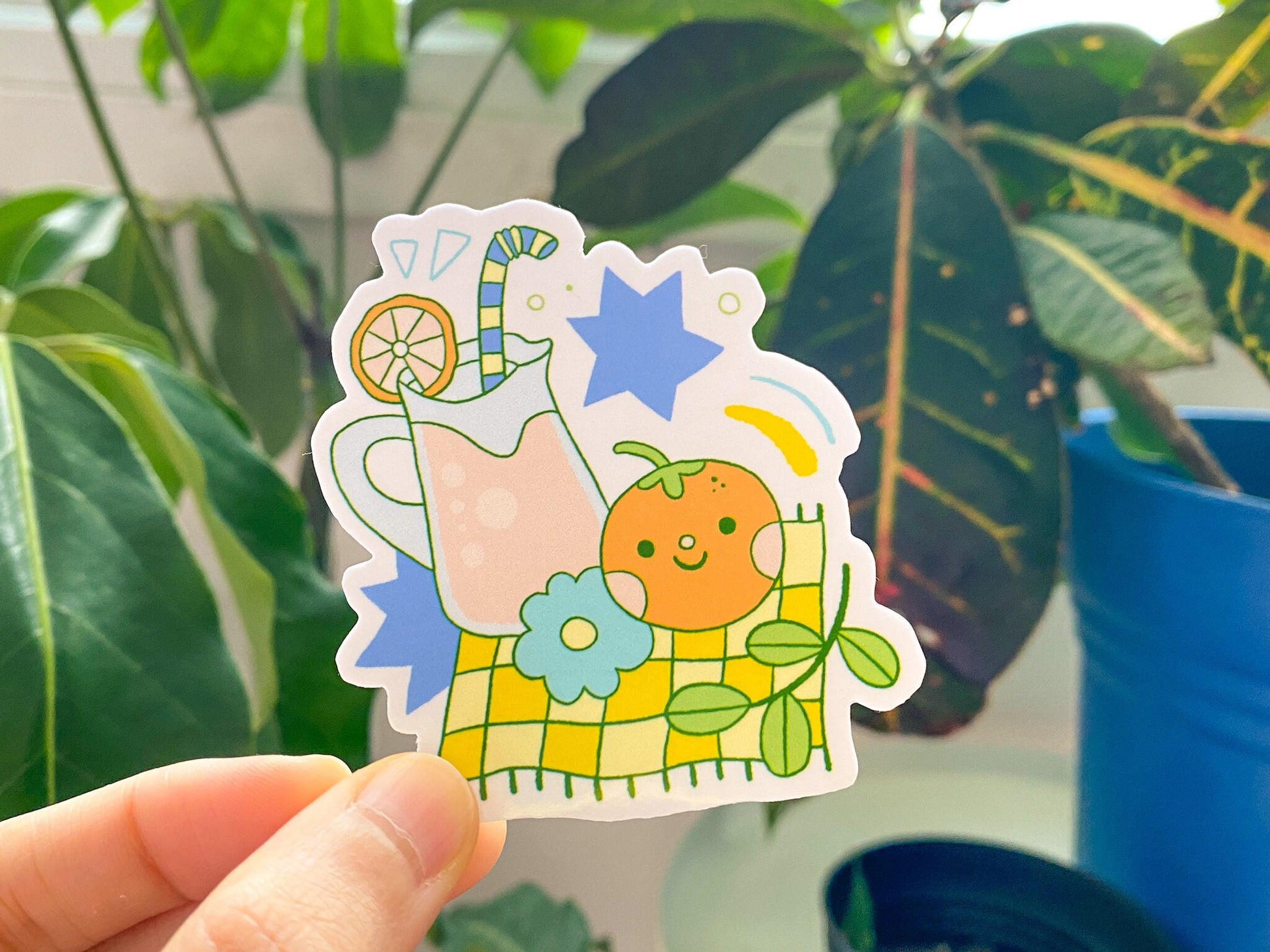 Sticker of a pitcher with lemonade, orange, and flowers held in front of green leaves.