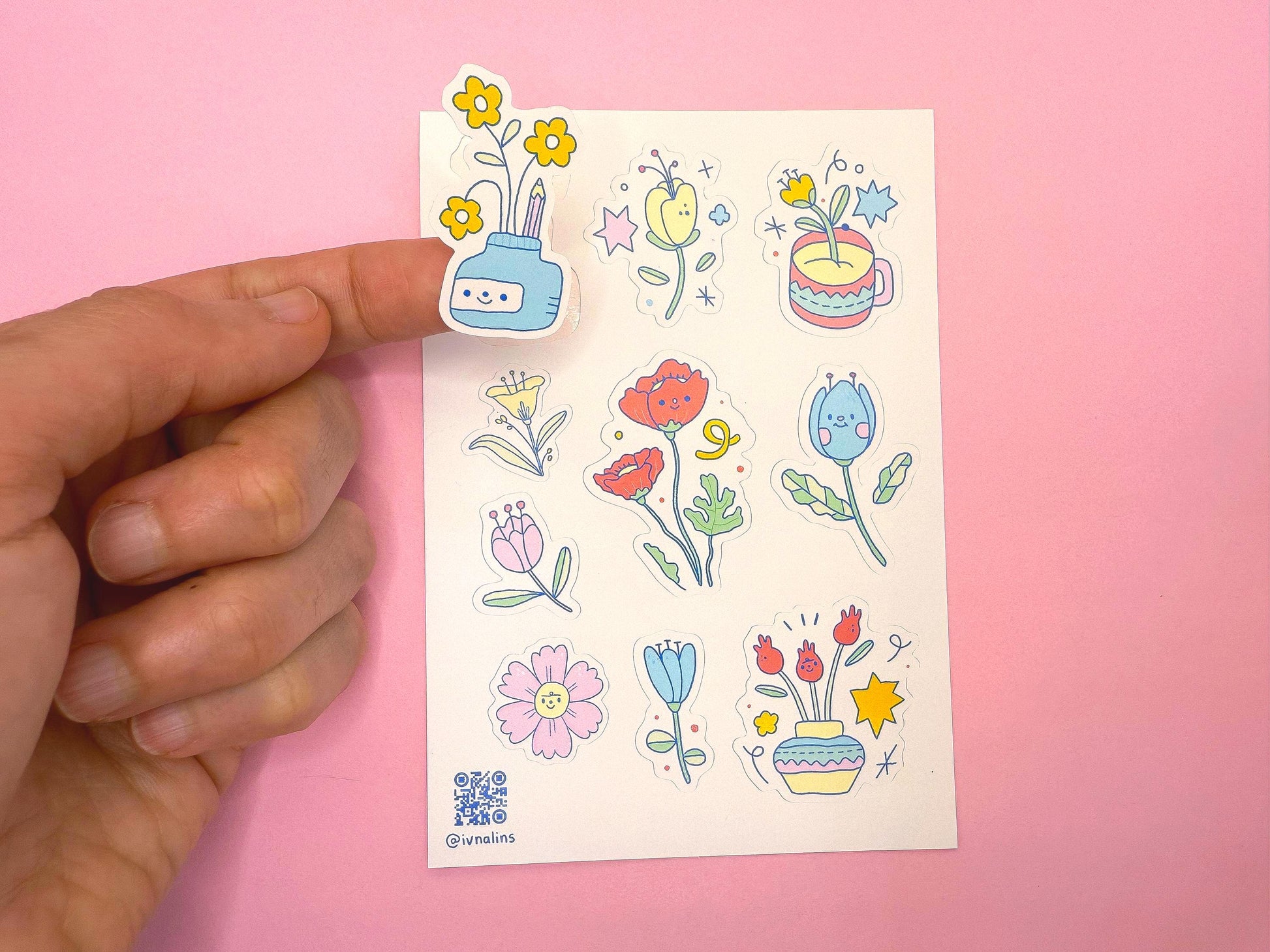 Sheet of colorful floral and plant stickers held by a hand against a pink background