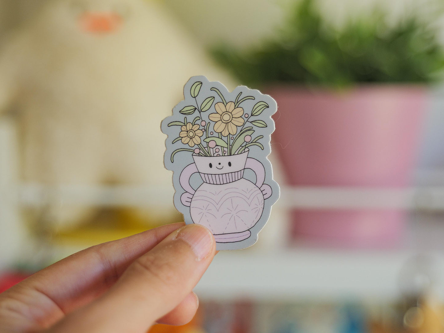 Hand holding a sticker of a cute vase with flowers against a blurred indoor background
