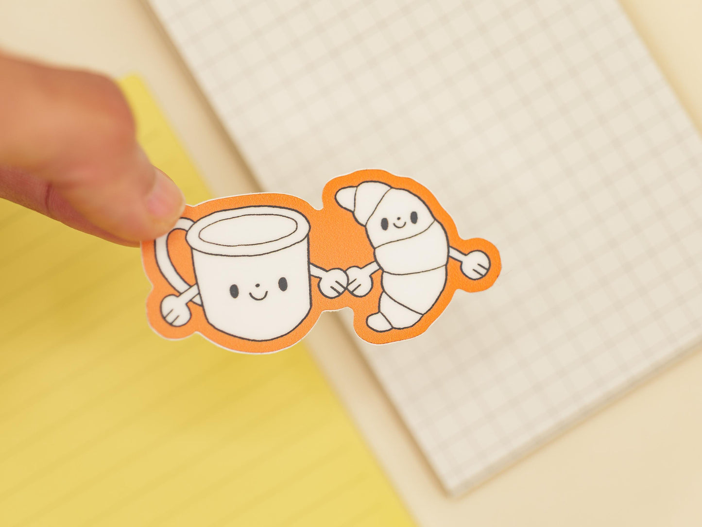 Two cartoon characters, a mug and a croissant, holding hands on a on a yellow background with a grid notebook.