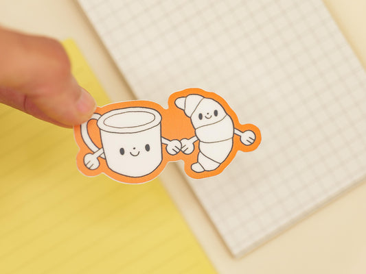 Two cartoon characters, a mug and a croissant, holding hands on a on a yellow background with a grid notebook.