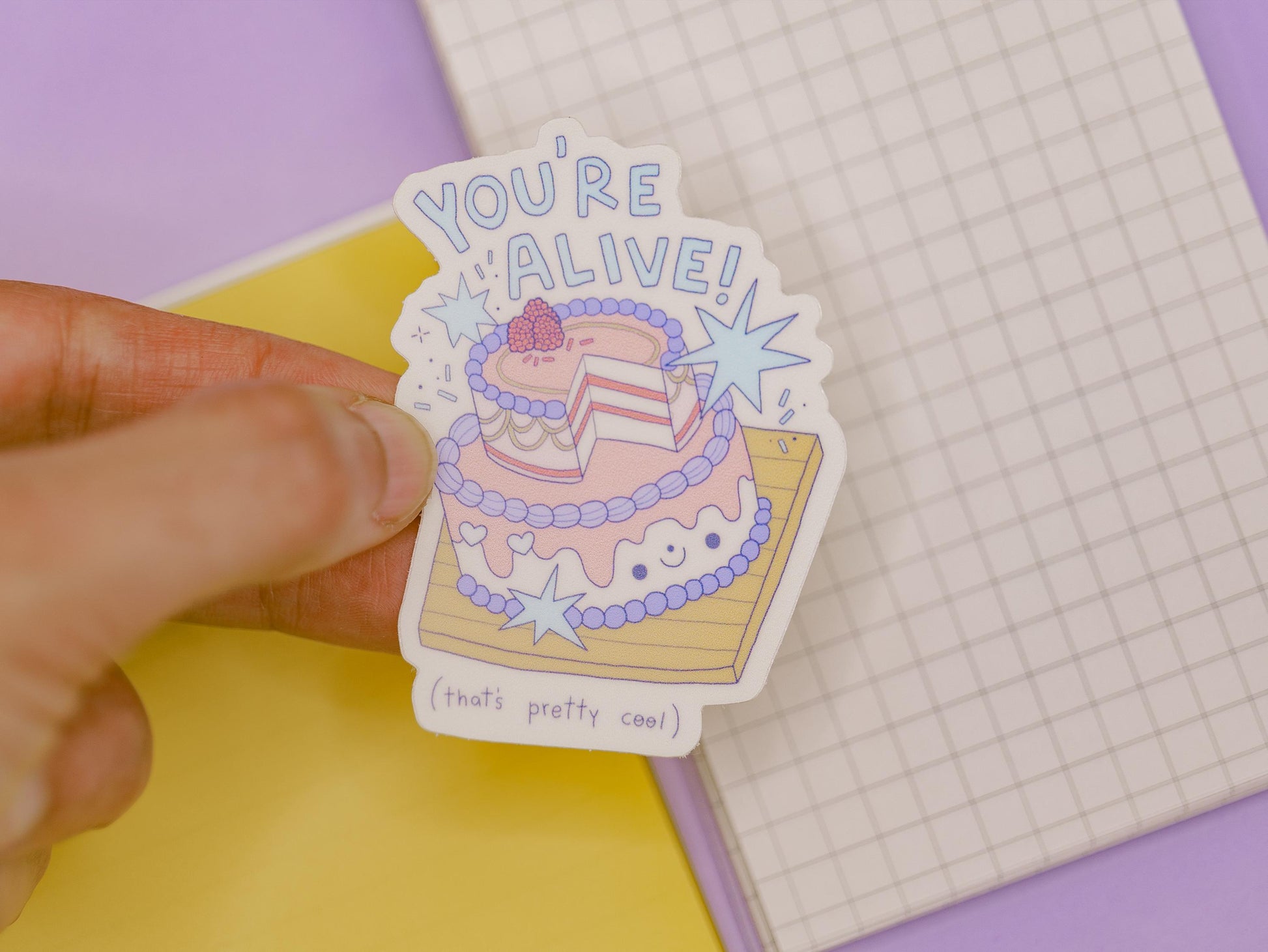 Sticker with a cake design and text held by a hand on a colorful background