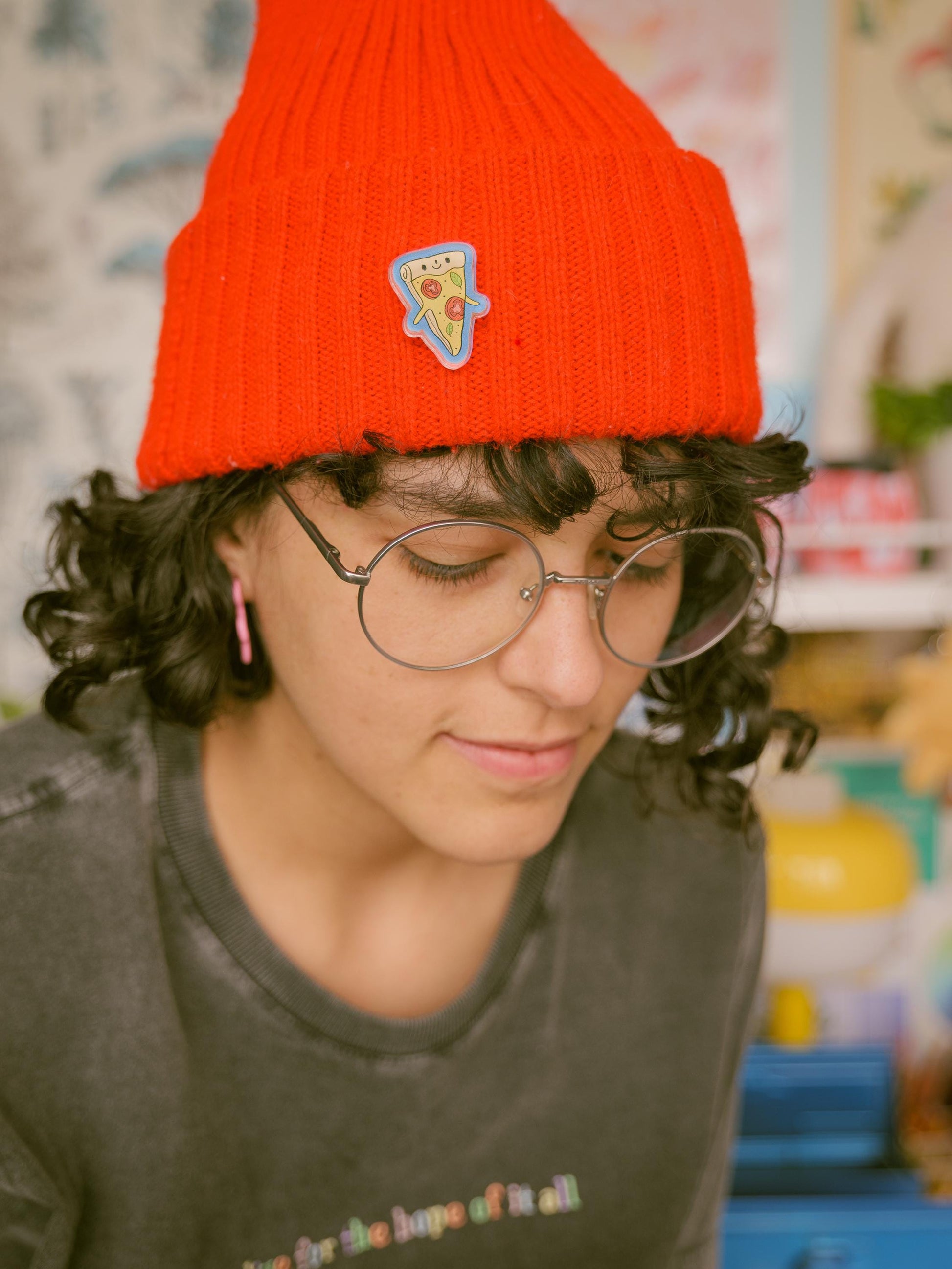 Person wearing a red beanie with a pizza pin and glasses, looking down.